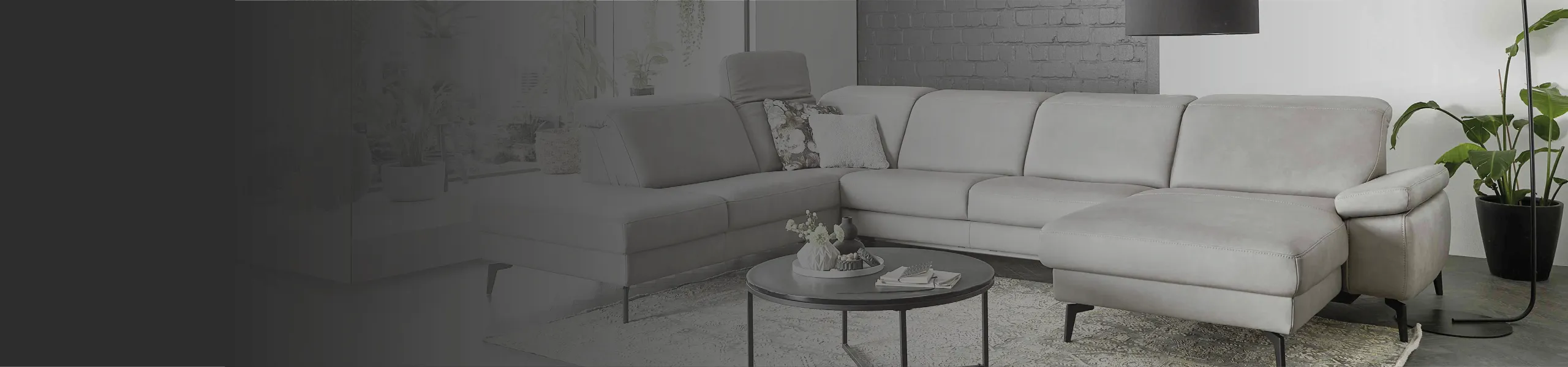 Stilvolles hellgraues Ecksofa mit verstellbaren Kopfstützen, gemütlichen Dekokissen und schwarzem Couchtisch im modernen Wohnzimmer mit Backsteinwand und Pflanzen. Perfekt für Wohnen mit Komfort und Design.